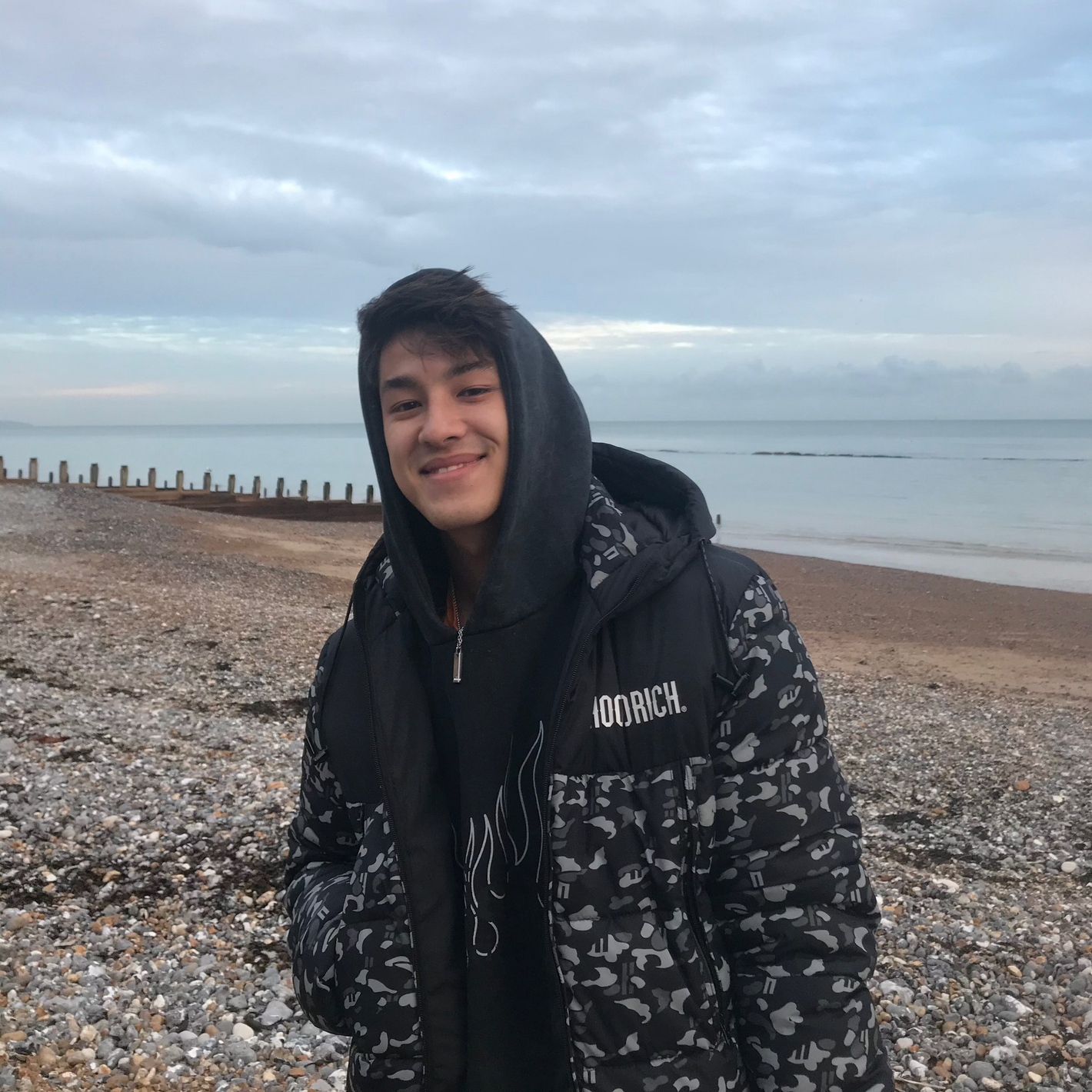 Young man smiling on a quiet pebble beach during a cloudy day, exploring a hidden coastal spot found using Sole Drift.