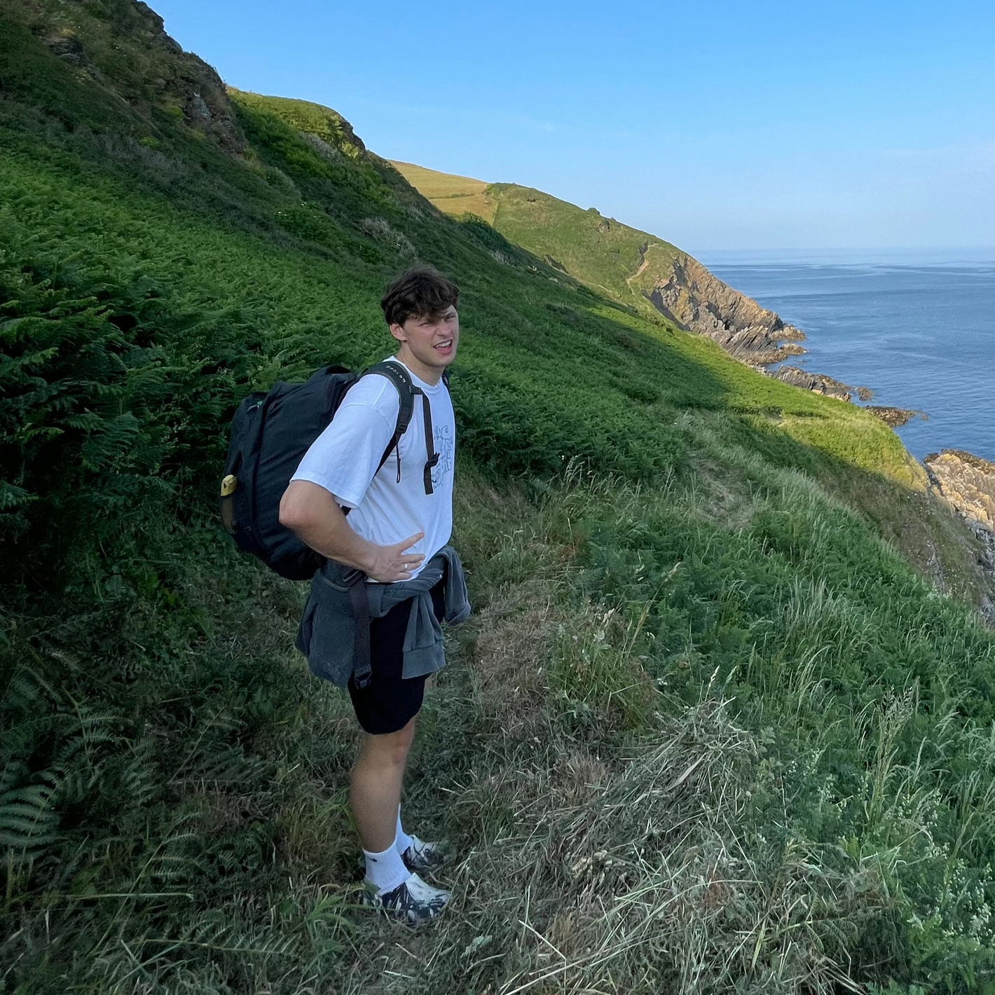 Male backpacker standing on a coastal hiking trail overlooking the ocean, exploring a remote nature spot found using the Sole Drift map.