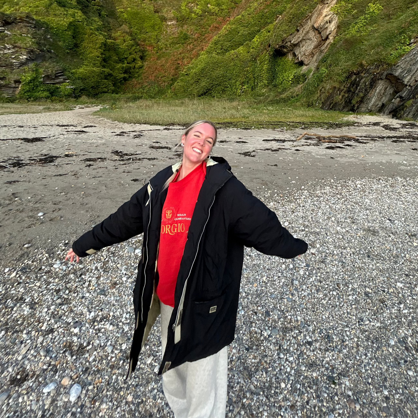 Smiling woman standing on a remote pebbled beach surrounded by cliffs and greenery, enjoying a hidden coastal spot discovered using Sole Drift.