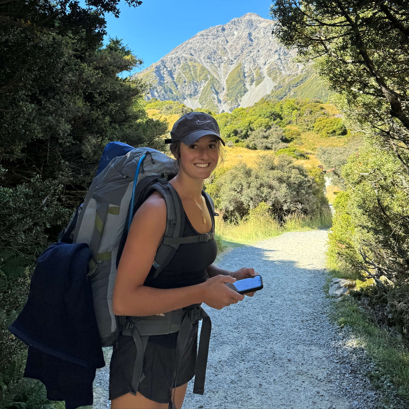 Female hiker smiling on a forested mountain trail, wearing a backpack and holding a phone with the Sole Drift map open, showcasing outdoor exploration in Australia.